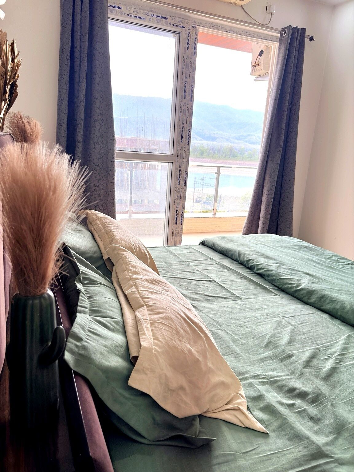A bed with green bedding, a vase with dried plants, and a window with curtains.