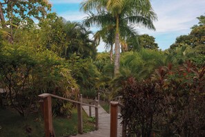 Eine Holztreppe führt durch einen üppigen Garten mit verschiedenen tropischen Pflanzen und einer Palme.
