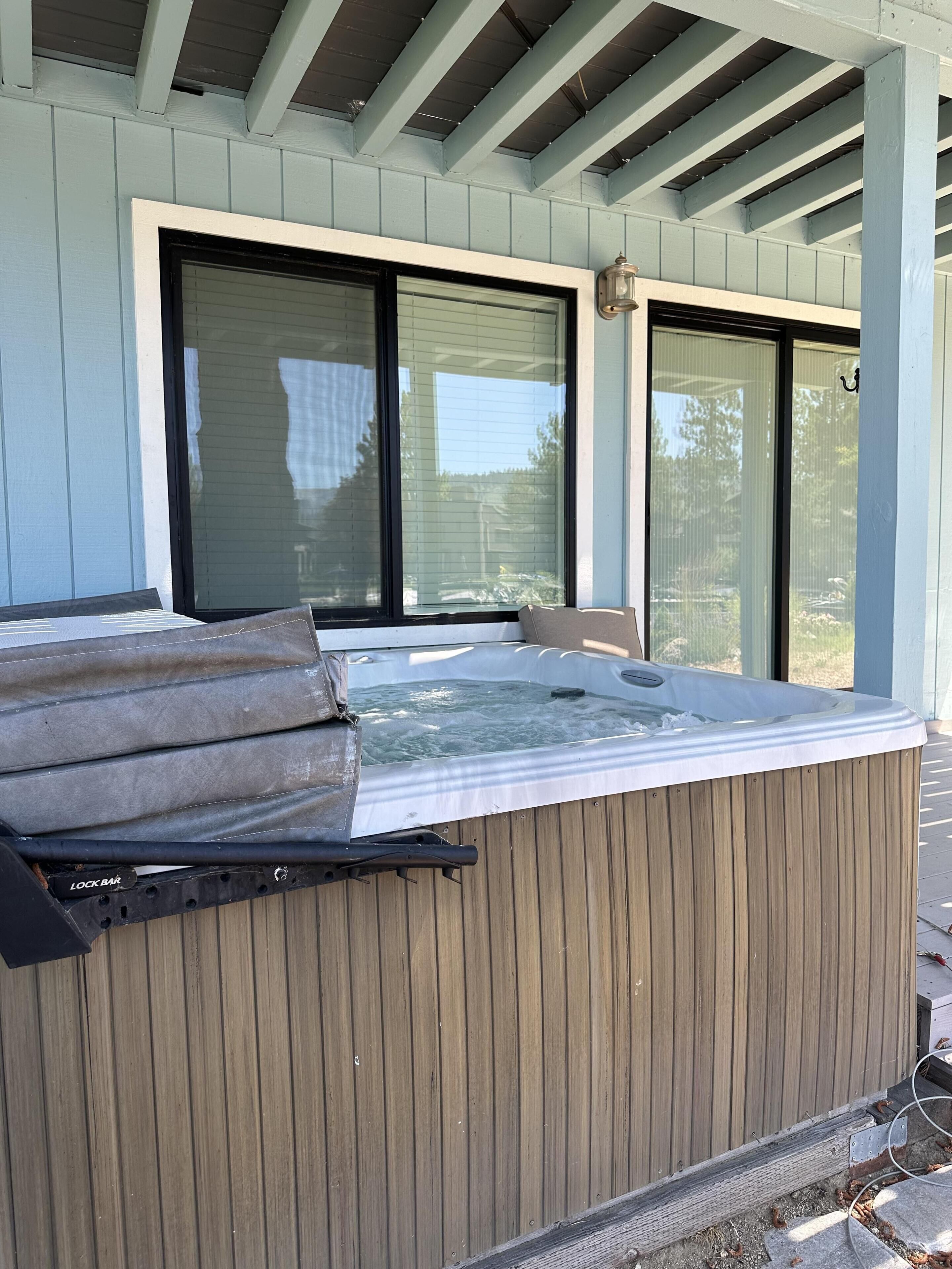 A hot tub with a wooden exterior, positioned under a covered patio area with large windows.
