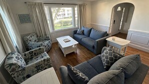 En stue med sofa, lænestol og sofabord, et vindue med gardiner, og et skilt på væggen.
