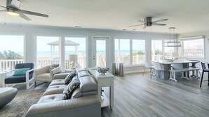 Een ruime woonkamer met een grote bank, eettafel en stoelen, een plafondventilator en uitzicht op het strand door grote ramen.