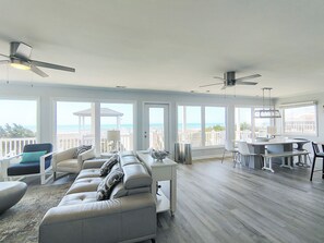 Ein geräumiger Wohnbereich mit einer großen Couch, Esstisch und Stühlen, einem Deckenventilator und Blick auf den Strand durch große Fenster.