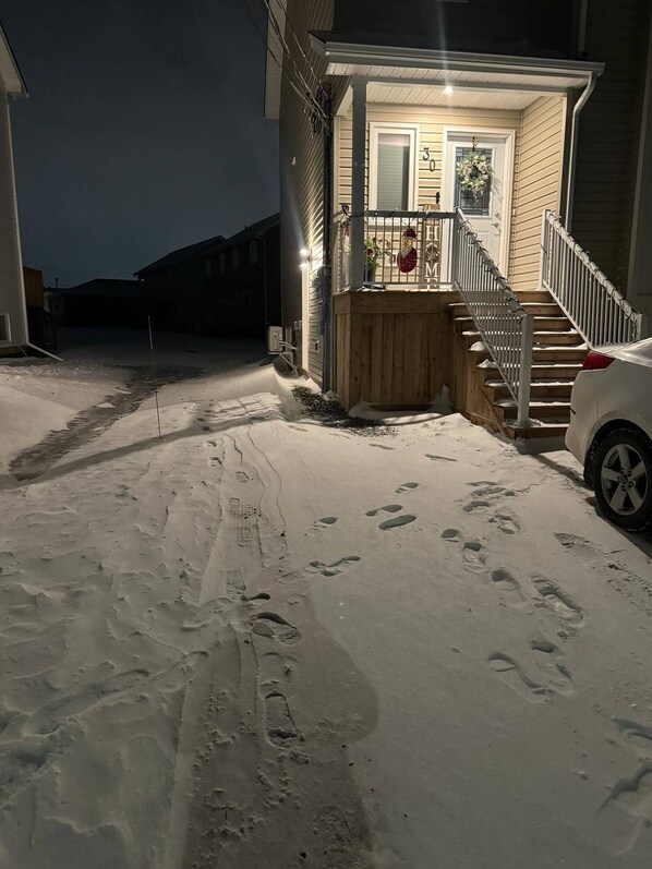 A snow-covered driveway leading to a house with a wooden deck and a staircase.