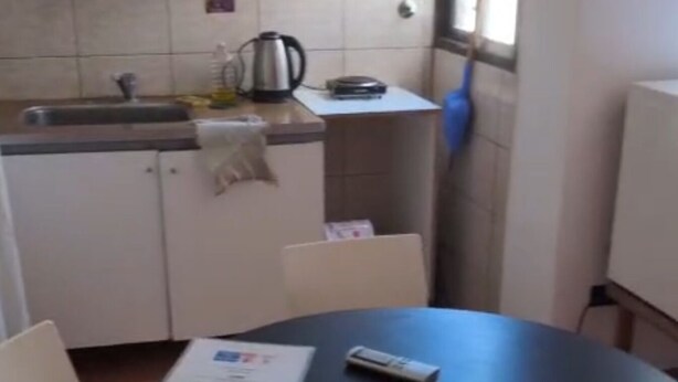 A small kitchen with white cabinets, a sink, and a window. There is a round table with chairs and a remote control on it.