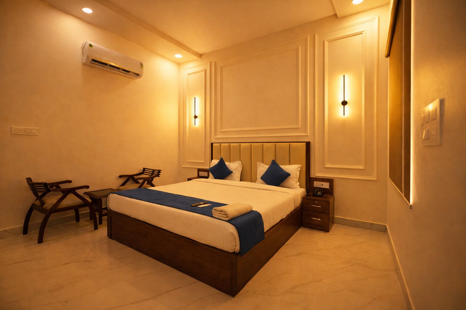 Une chambre d’hôtel comprenant un lit, des tables de chevet, un bureau avec une chaise, un appareil de climatisation et des luminaires fixés au mur.