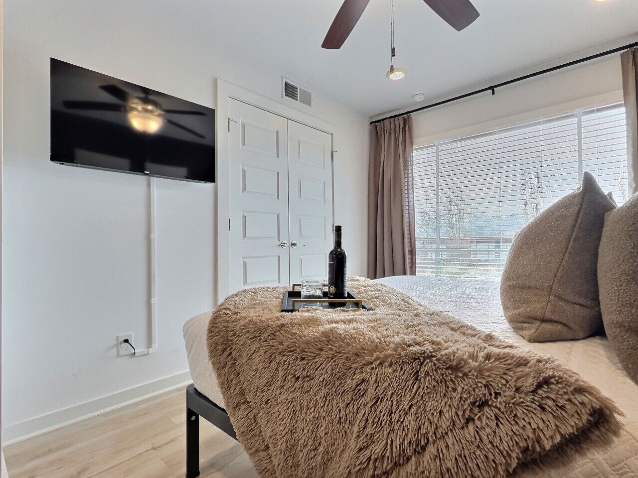 A modern bedroom with a bed, a television mounted on the wall, a ceiling fan, and a window with curtains.