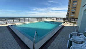 Une piscine rectangulaire bordée d’une rambarde métallique, entourée d’une terrasse dallée avec des chaises longues, d’où l’on peut admirer une plage et un immeuble de grande hauteur.