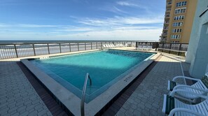 Ein rechteckiger Pool mit Metallgeländer, umgeben von einer gepflasterten Liegefläche mit Liegestühlen, mit Blick auf einen Strand und ein Hochhaus.
