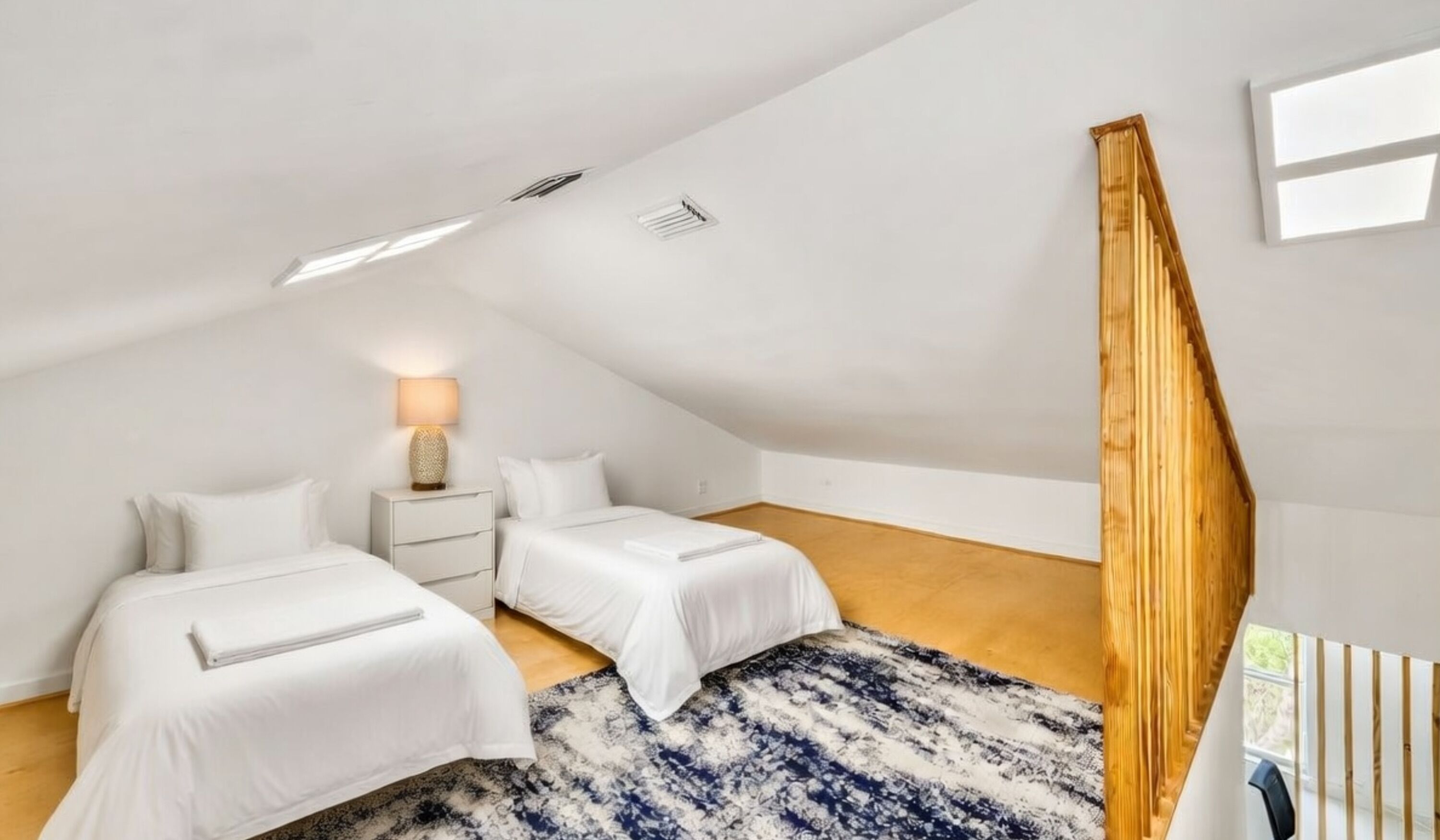 Ein modernes, helles Schlafzimmer im Dachgeschoss mit zwei Betten, einem Nachttisch, einer Lampe und einer Holztreppe.