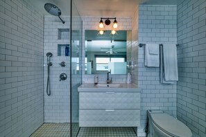Un baño moderno con paredes de azulejos blancos, cerramiento de ducha de vidrio, un lavabo blanco con espejo superior y un inodoro.