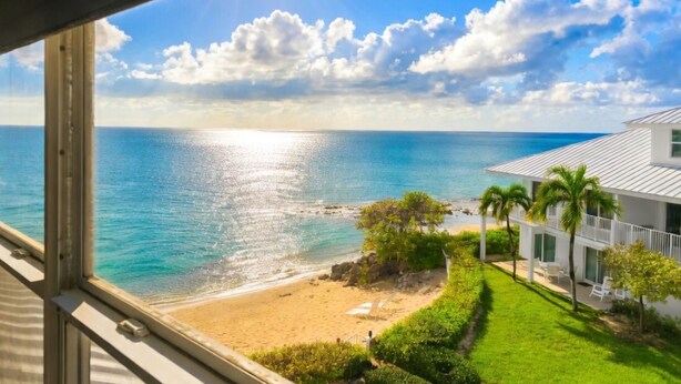 A beach view from a window, including a sandy beach, ocean, and a white building with a balcony. - 7 Mile Beach | Soft Sand, Epic Sunset Views & Shipwreck Snorkel (Grand Cayman)