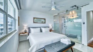 A modern hotel room with a large bed, a ceiling fan, a glass-enclosed shower, and a view of the city. - 7 Mile Beach | Soft Sand, Epic Sunset Views & Shipwreck Snorkel (Grand Cayman)