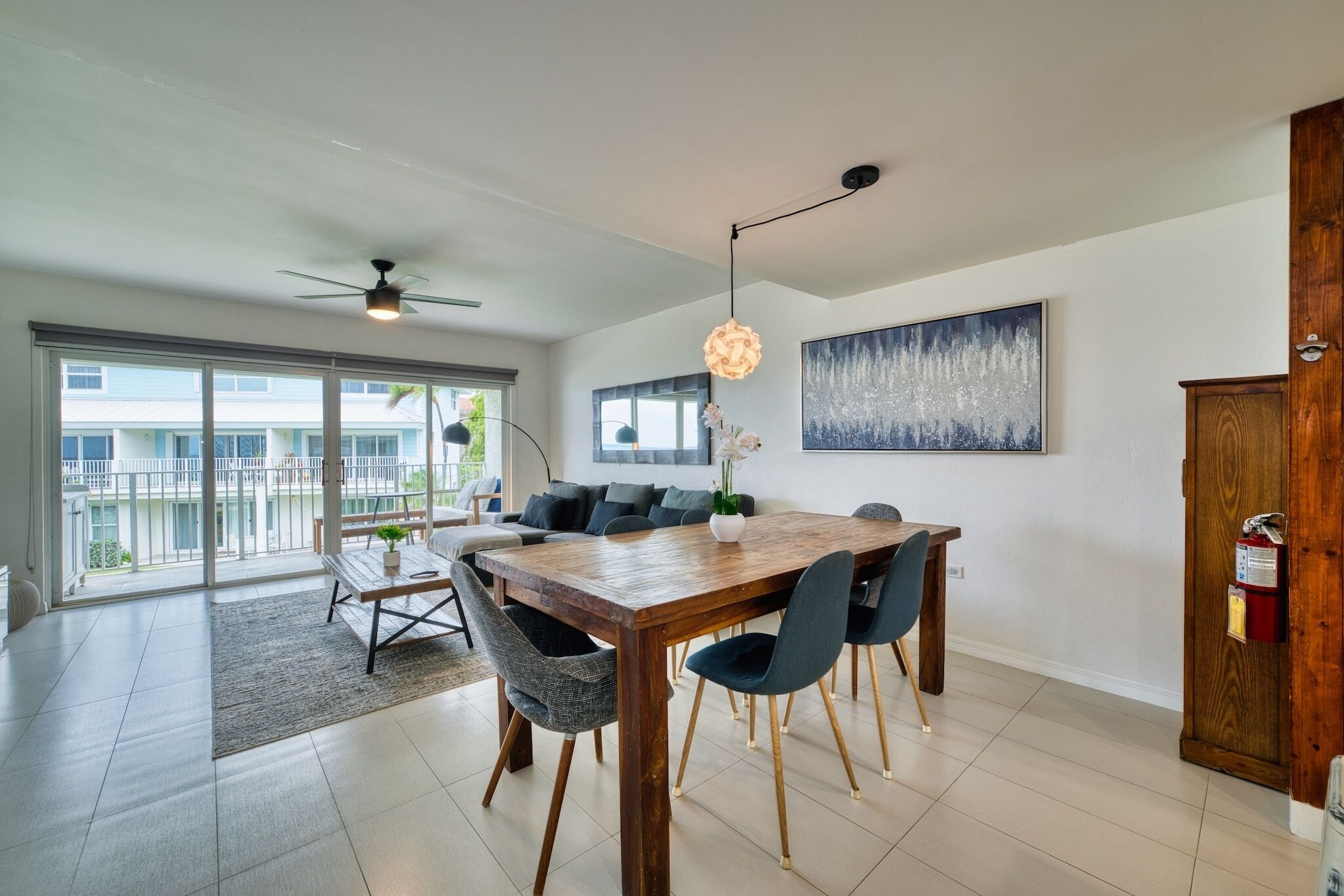 A modern living room with a wooden dining table, dark chairs, a ceiling fan, and a large window.