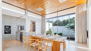 A modern kitchen with built-in appliances, a dining area with a long table and chairs, and large windows offering a view of the outdoors. - New Prime Location Near Boat Avenue V306 (Choeng Thale)