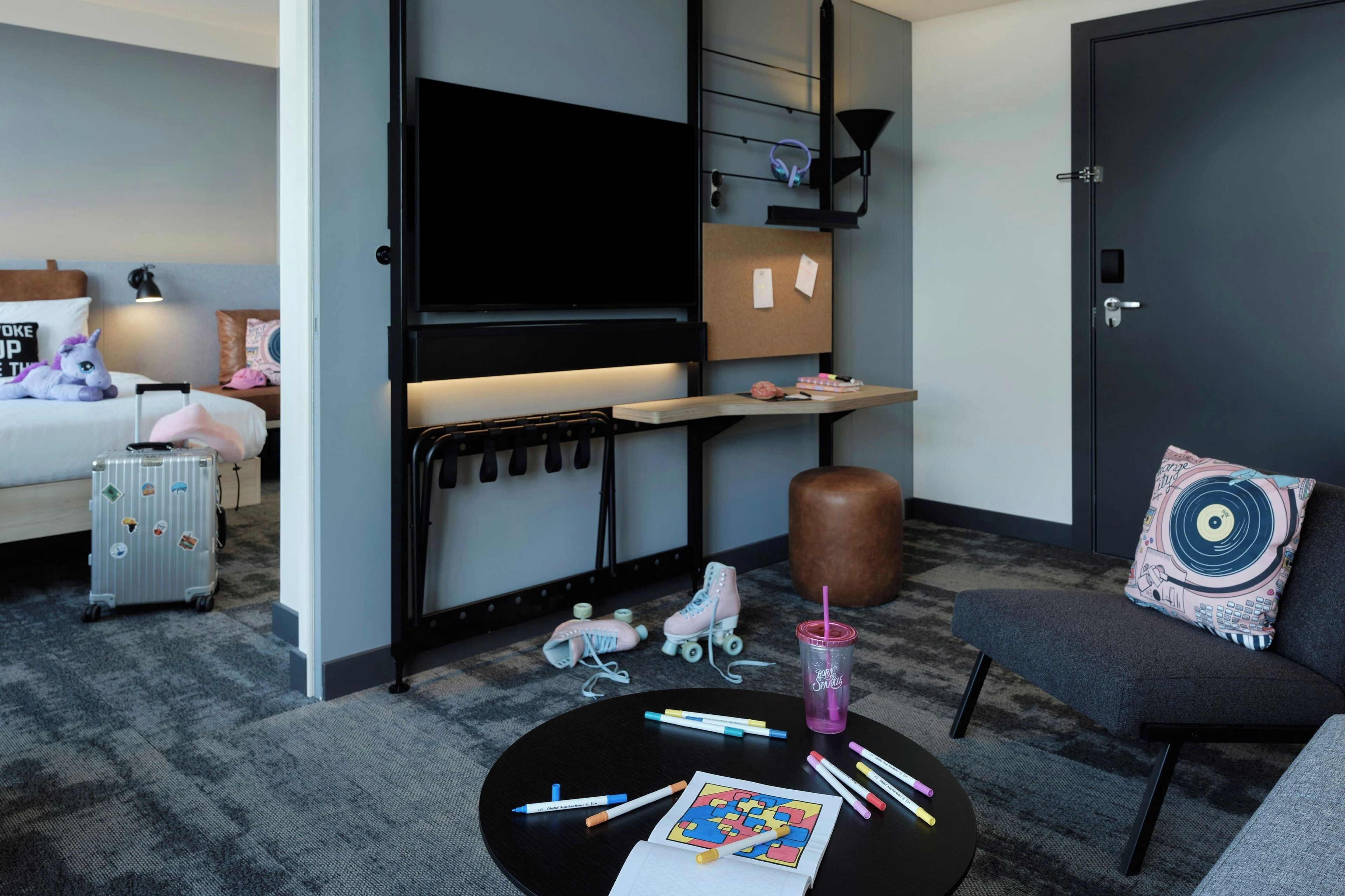 Habitación de hotel con cama, escritorio, televisor, zona de estar y una pequeña mesa con marcadores y una bebida.