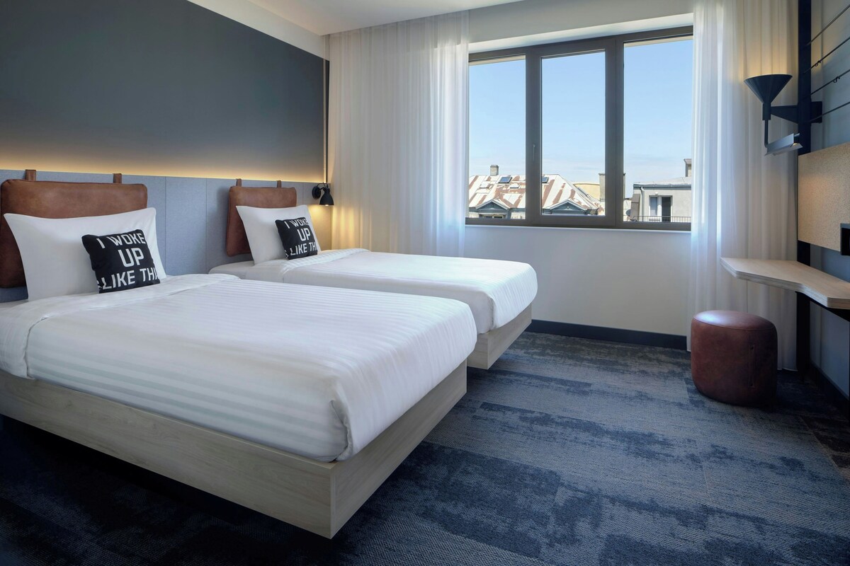Une chambre d’hôtel avec deux lits, une grande fenêtre donnant sur la ville, un petit pouf rond et une étagère fixée au mur.