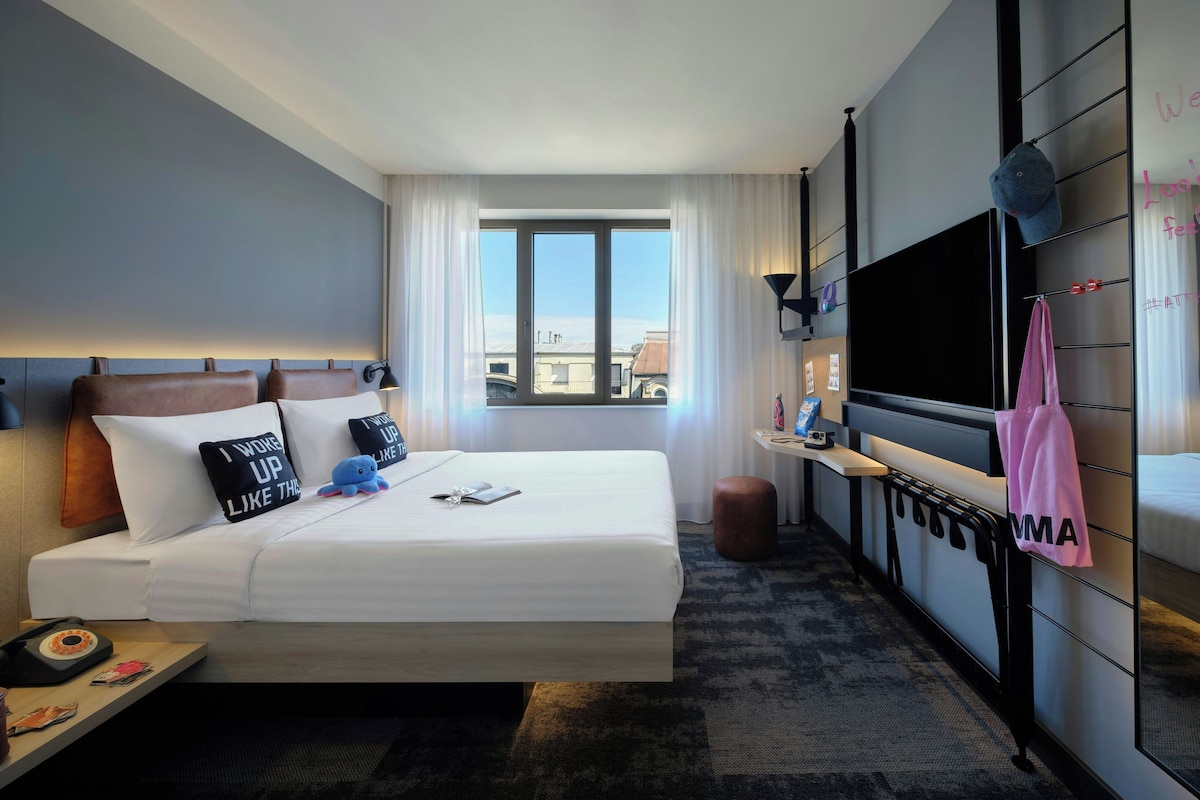 Une chambre d’hôtel équipée d’un lit, d’un bureau, d’une télévision et d’une fenêtre donnant sur la ville.