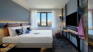 Une chambre d’hôtel équipée d’un lit, d’une télévision, d’un bureau et d’une fenêtre donnant sur des immeubles.