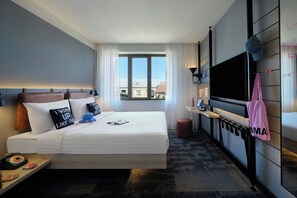 A hotel room with a bed, a TV, a desk, and a window with a view of buildings.