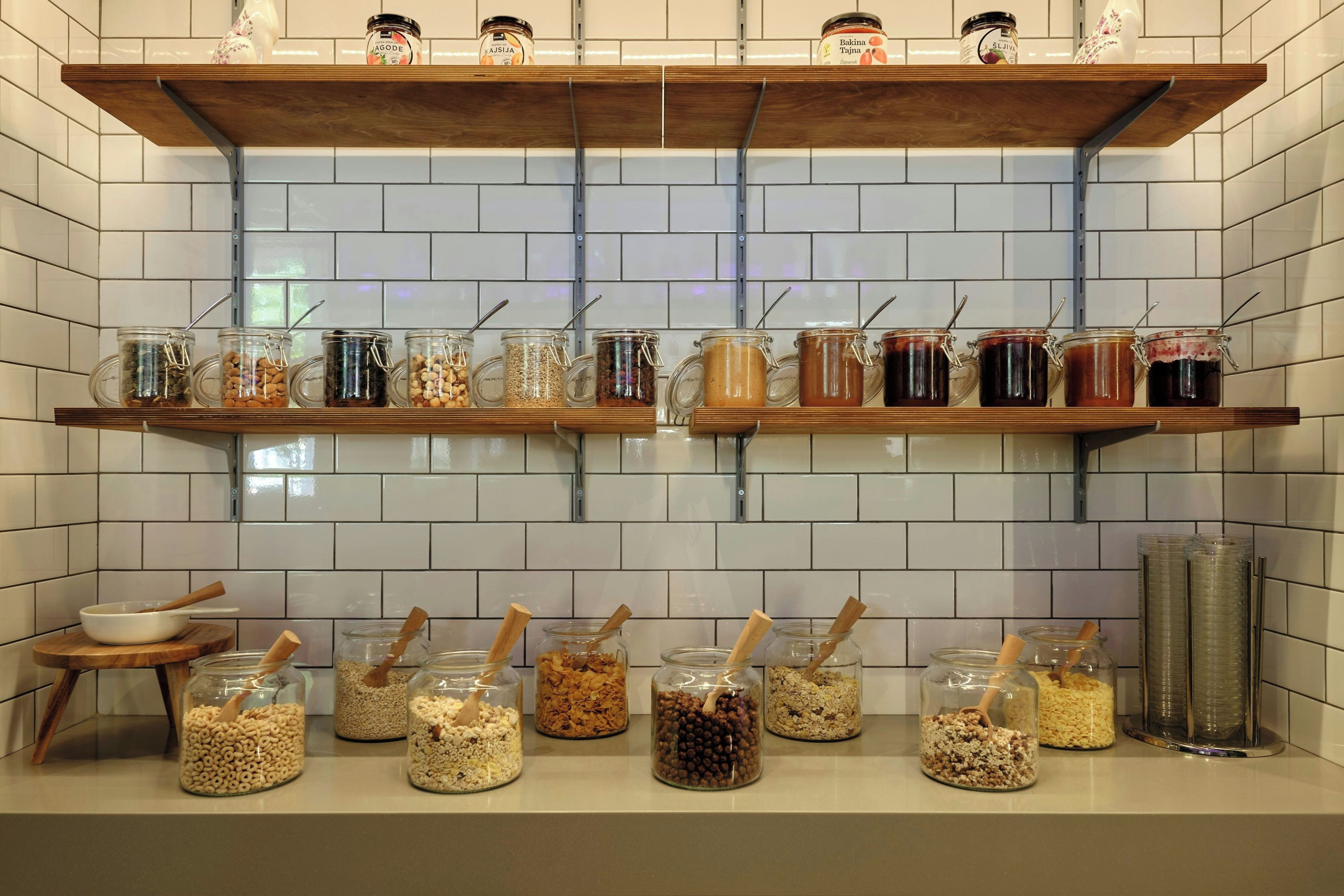 Una cocina con dos repisas de madera que sostienen frascos con alimentos y una encimera con frascos de cereales y nueces.