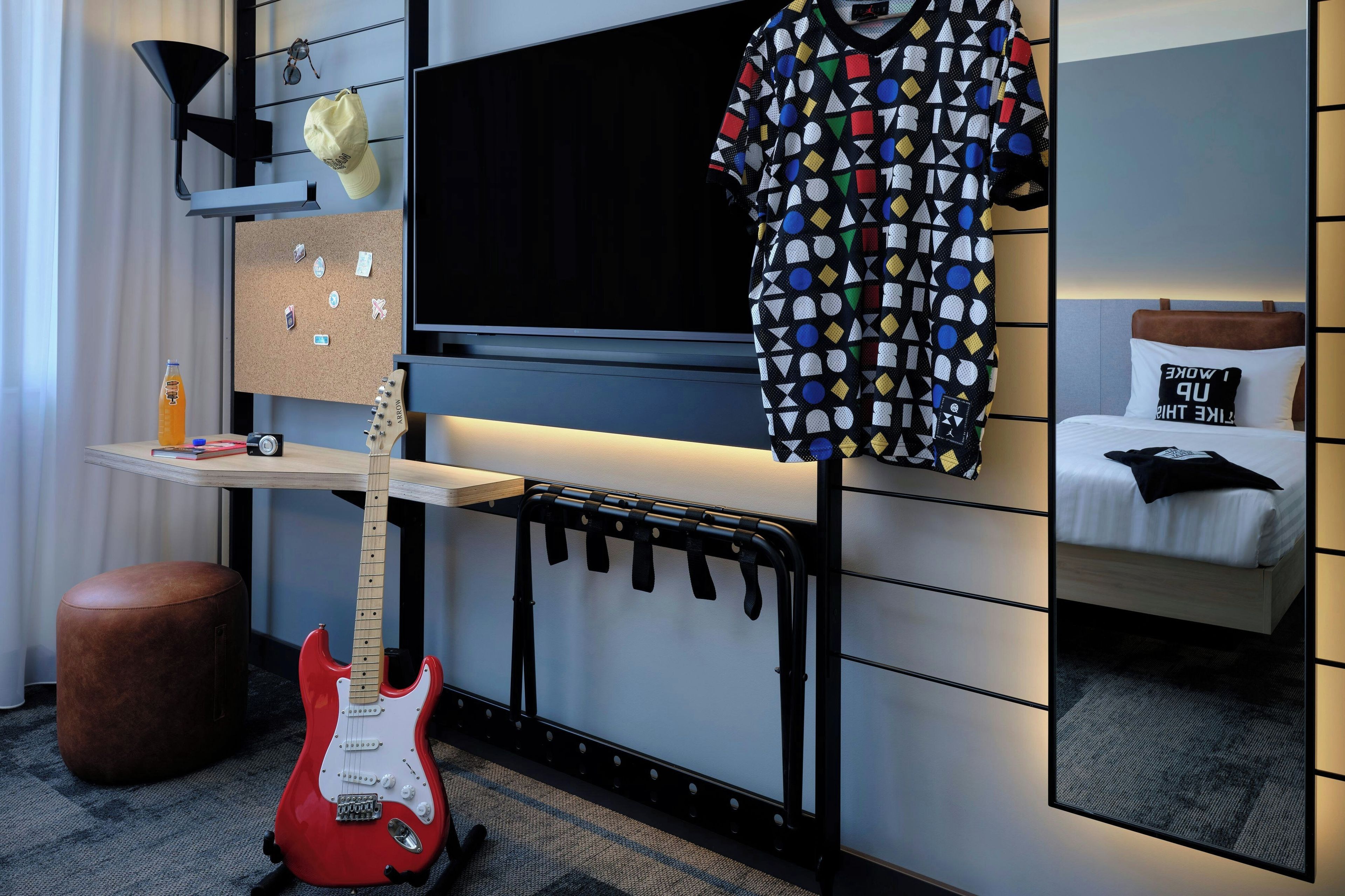 Une chambre d’hôtel équipée d’un lit, d’une télévision, d’une guitare, d’une étagère fixée au mur et d’un panneau d’affichage où divers objets sont accrochés.