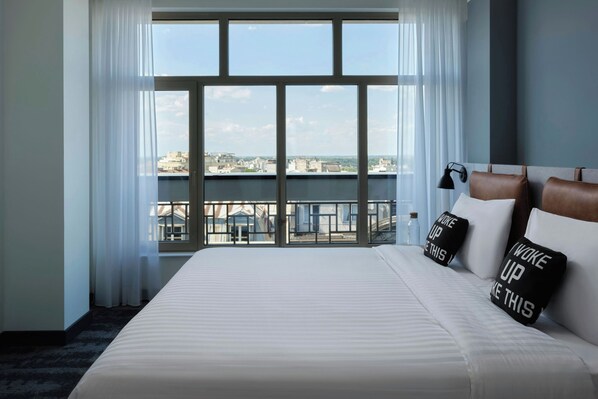 Habitación de hotel con una cama grande, vistas de la ciudad a través de ventanales del suelo al techo y un balcón con barandilla.