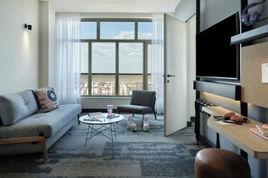 Una sala de estar moderna con un sofá gris, una mesita redonda pequeña, una silla y un ventanal grande con cortinas.