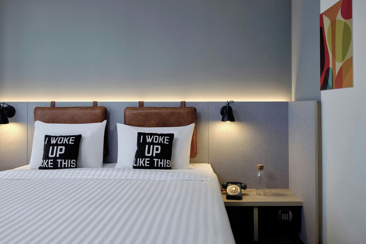 Une chambre d’hôtel comprenant un lit, deux oreillers ornés d’inscriptions motivantes, une table de chevet avec une lampe, un téléphone et un élément décoratif mural.
