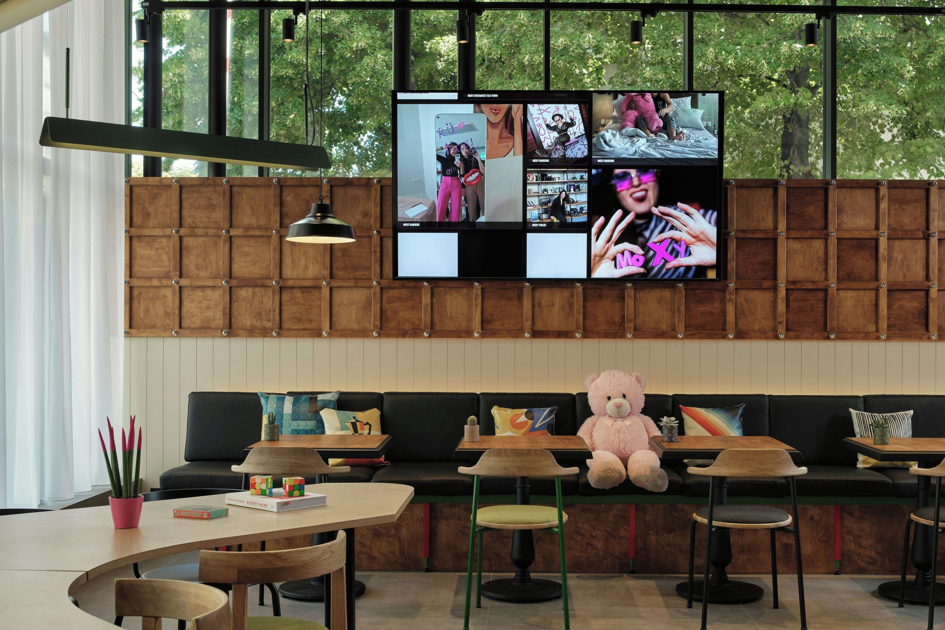 A modern interior with a large TV screen displaying multiple images, a sofa, a teddy bear, and a dining area with wooden tables and chairs.