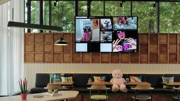 A modern interior with a large TV screen displaying multiple images, a sofa, a teddy bear, and a dining area with wooden tables and chairs.