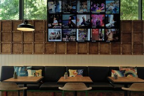 Un interior moderno con un televisor de pared grande mostrando varias escenas, un panel de pared de madera, un sofá negro con cojines de colores y una lámpara colgante.