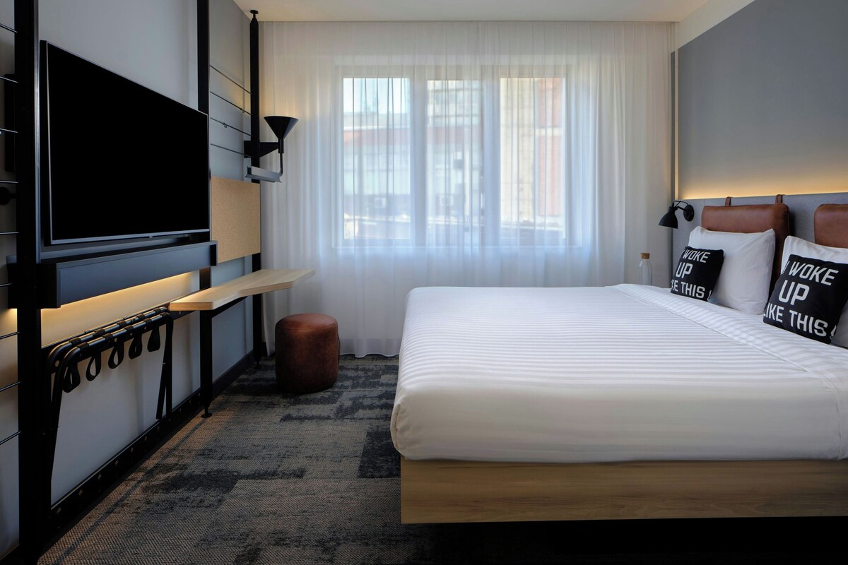 Habitación de hotel con una cama grande, un televisor de pantalla plana montado en la pared, una mesita pequeña y una ventana con cortinas translúcidas.