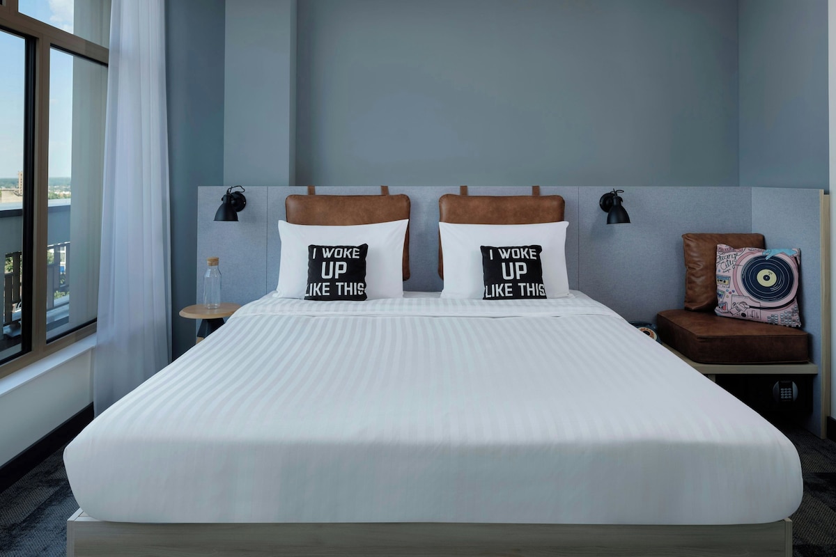 A hotel room with a large bed, two pillows with text, a nightstand, a lamp, and a view of the city.