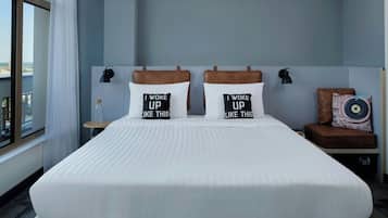 Habitación de hotel con una cama grande, dos almohadas con texto, una mesita de noche, una lámpara y vista a la ciudad.
