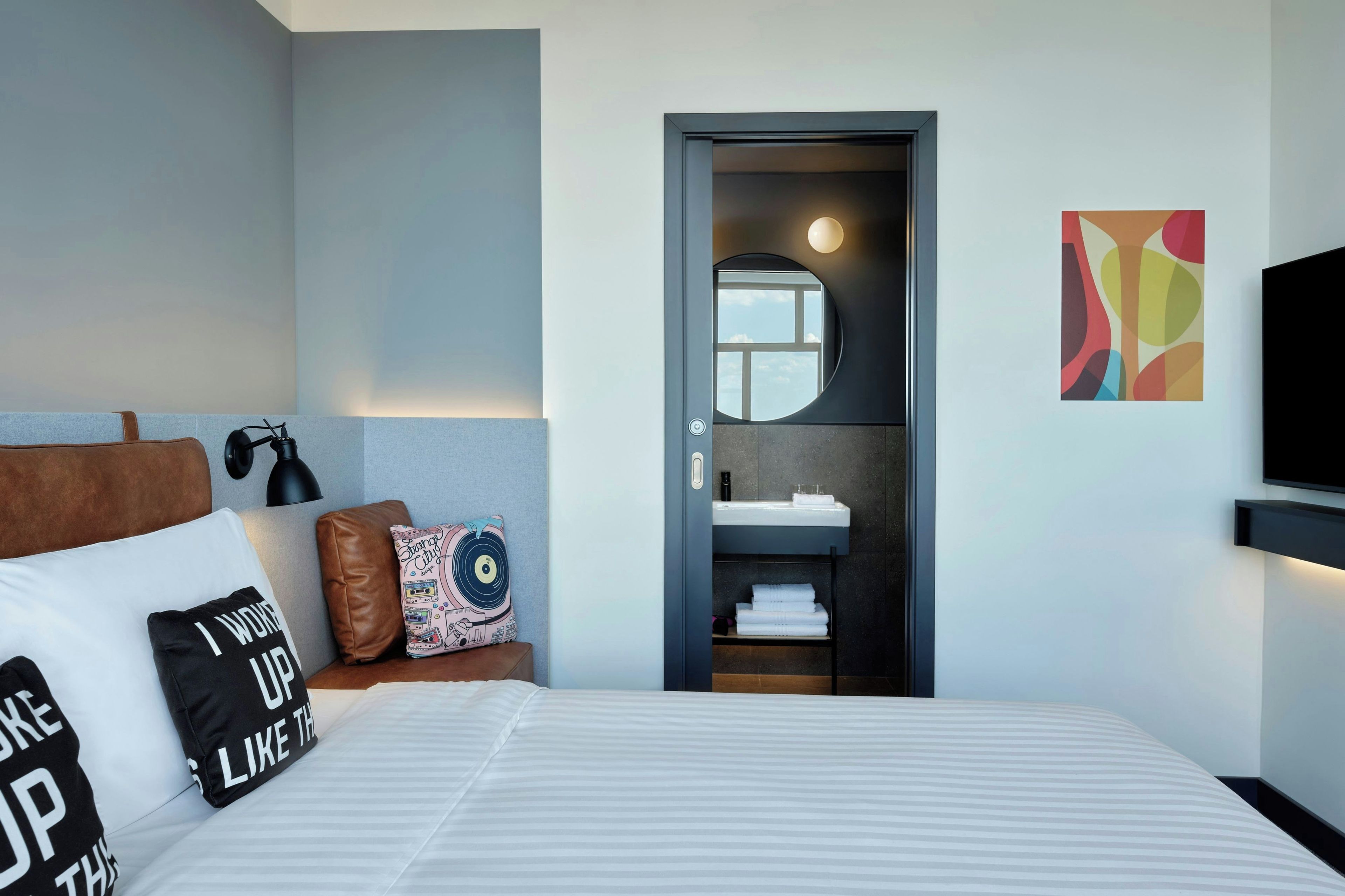 Ein modernes Hotelzimmer mit Bett, Nachttisch, Wand-Fernseher, Bad mit Waschbecken und Spiegel sowie einem farbenfrohen abstrakten Gemälde.