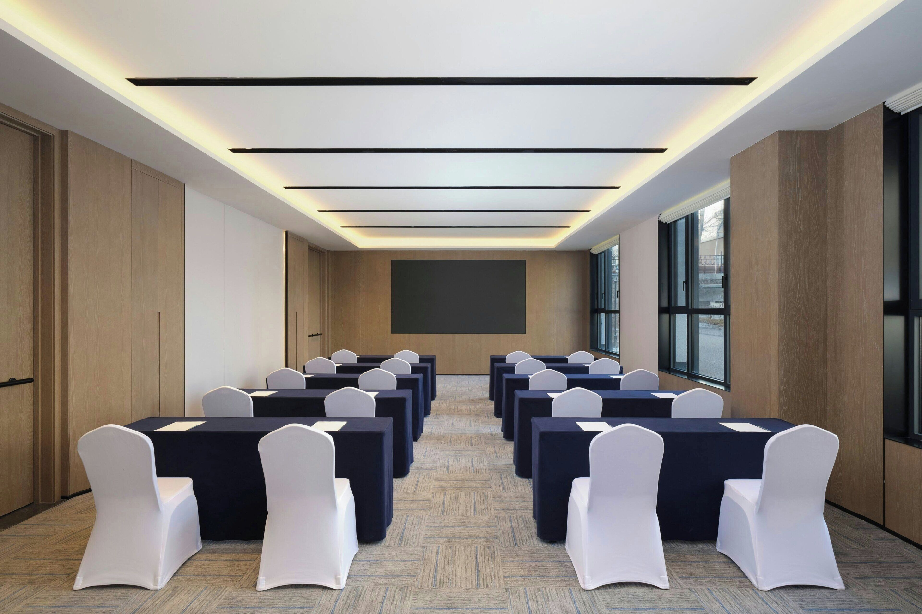 Un salón de conferencias con filas de mesas y sillas, una pantalla grande y un diseño de techo moderno.