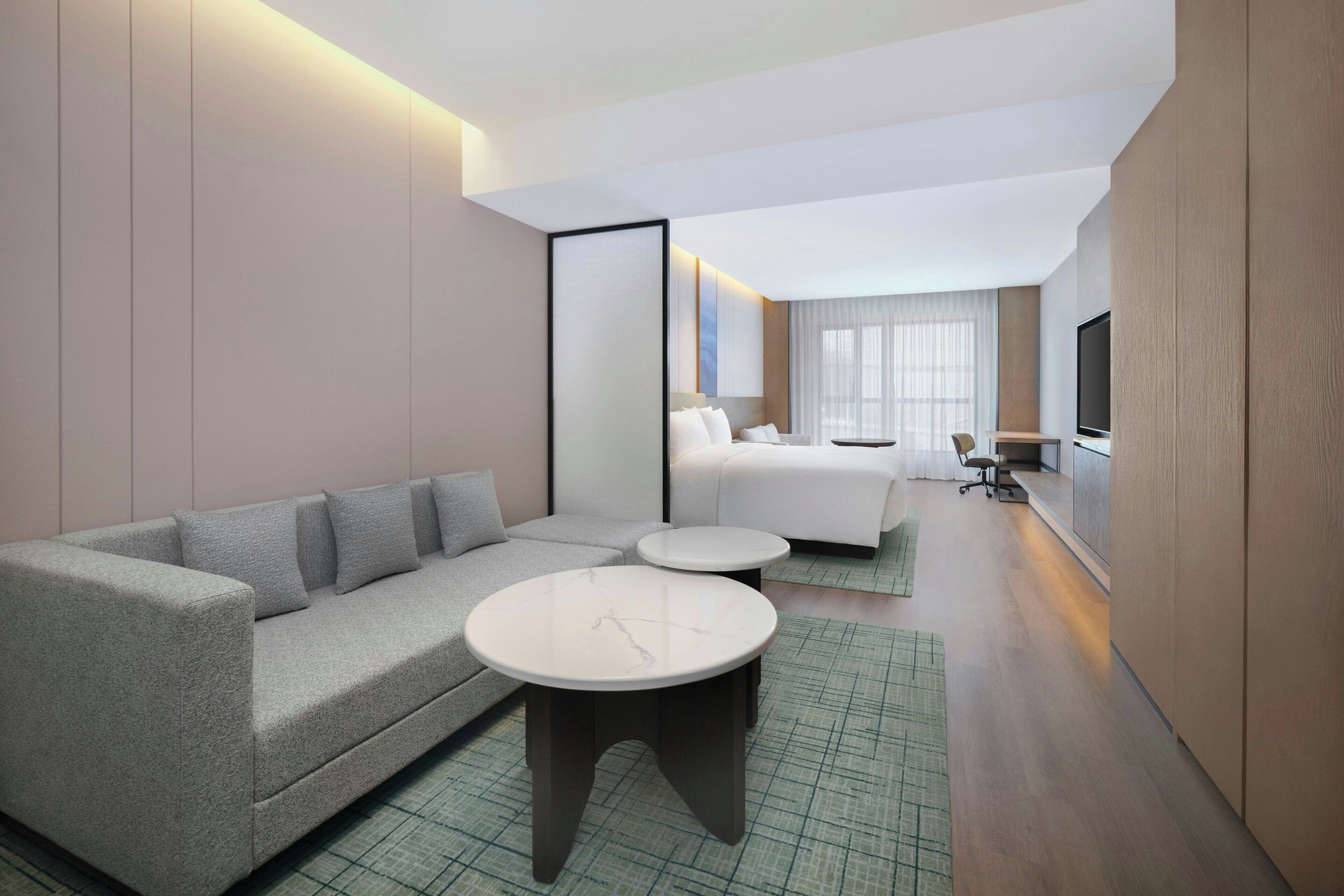 A modern hotel room with a grey sofa, two round tables, a bed, a TV, and a large window.