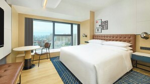 Une chambre d’hôtel avec un grand lit, un bureau, une chaise et une vue sur la ville par une grande fenêtre.
