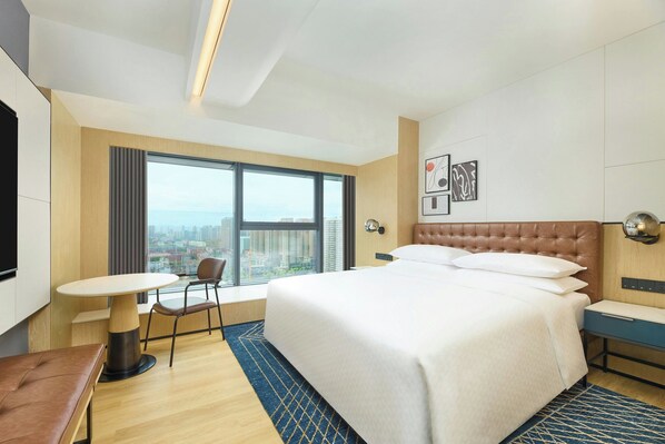 Ein Hotelzimmer mit einem großen Bett, einem Schreibtisch, einem Stuhl und Blick auf die Stadt durch ein großes Fenster.