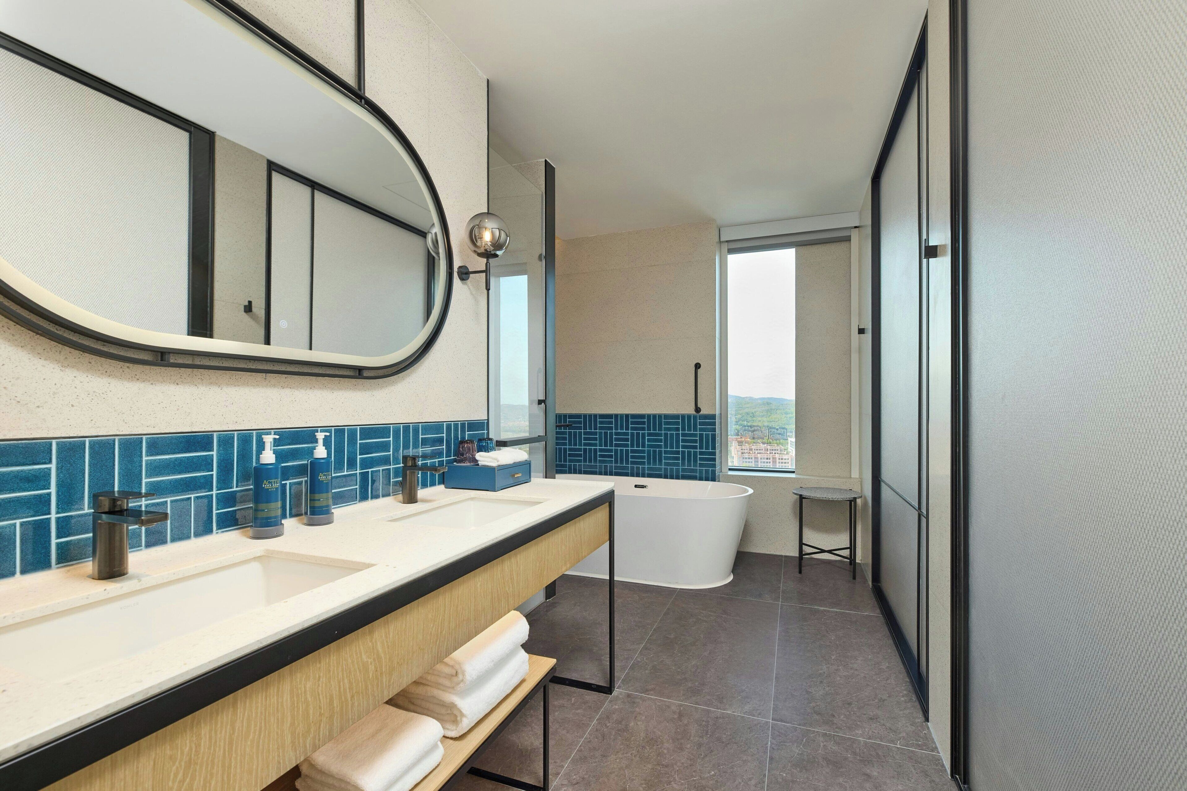 A modern bathroom with a large mirror, dual sinks, a bathtub, and a view through a window.