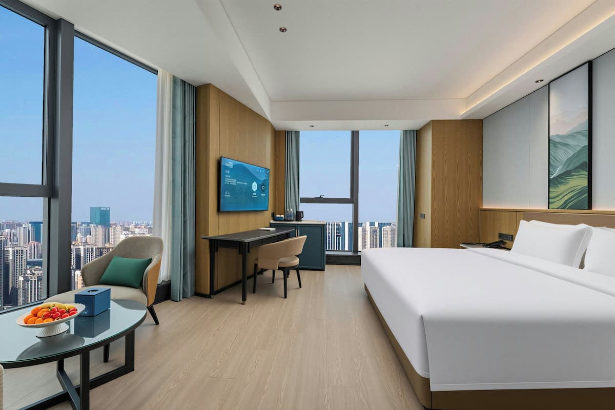 A modern hotel room with a large bed, a desk, a chair, a small table with fruit, a TV, and a city view.