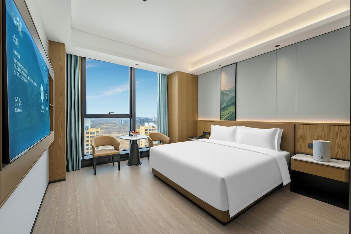 A modern hotel room with a large bed, a desk, a chair, a small table, and a large window offering a city view.