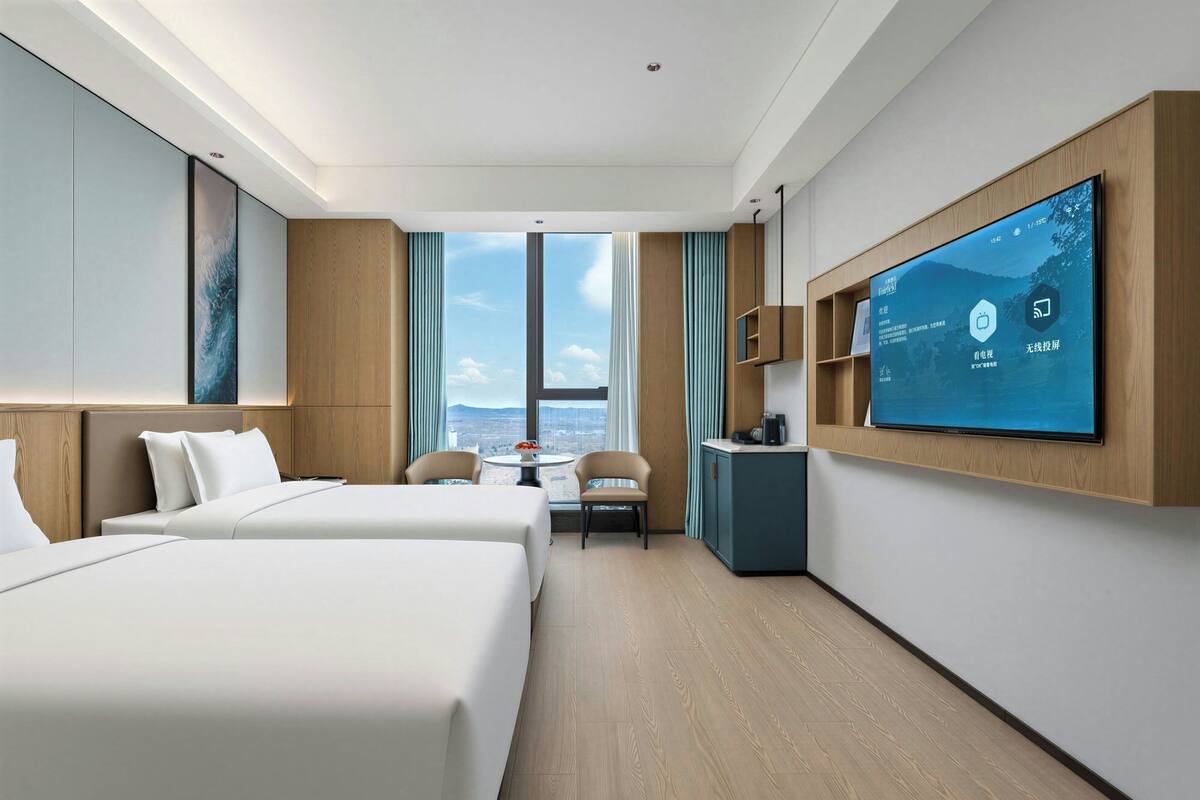 A hotel room with two beds, a desk, a TV, and a view of the city.