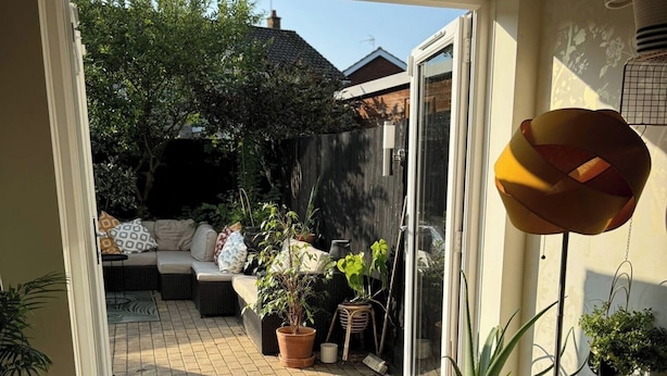 Une pièce dotée d’une porte-fenêtre coulissante donnant sur un patio extérieur aménagé avec un canapé, des plantes en pot et une lampe en métal.