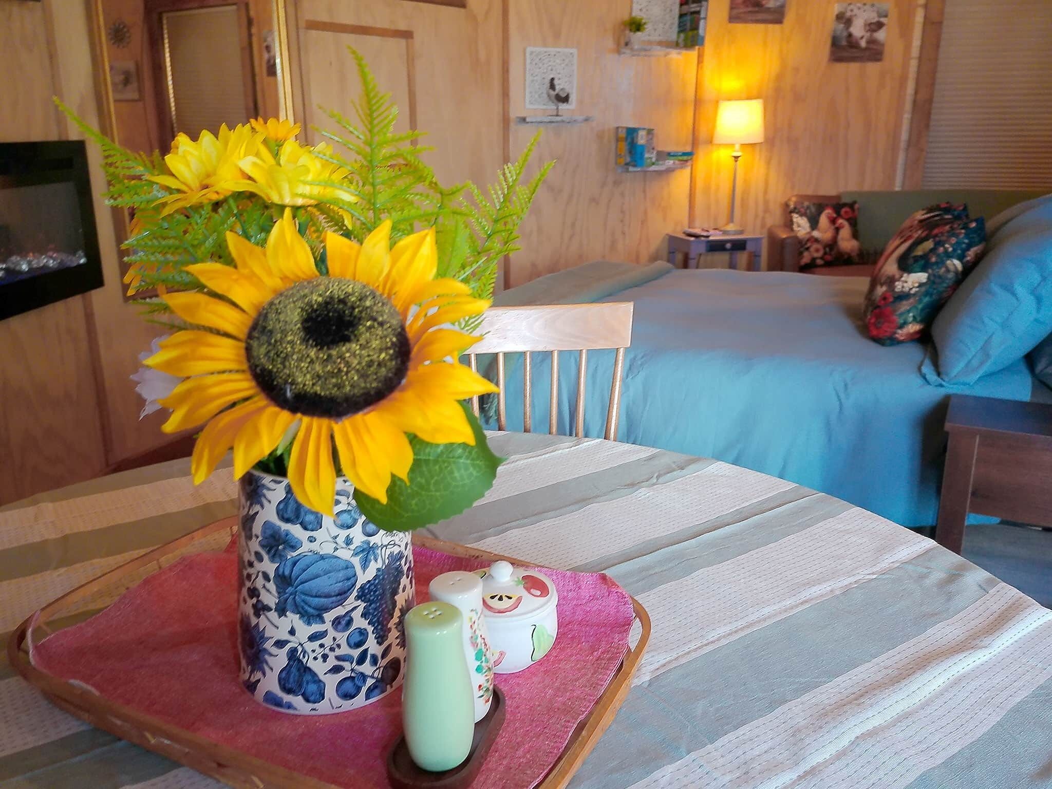 Une pièce avec un lit, une table sur laquelle est posé un vase rempli de tournesols, et une lampe.