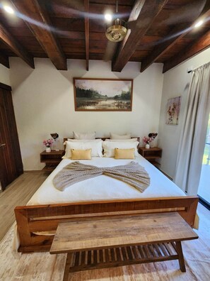 A bedroom with a wooden bed, a bench, a painting above the bed, and a nightstand with a vase. - Canary Suite - Brisal Mountain Retreat (Jarabacoa)