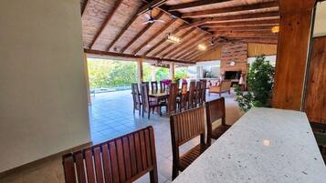 A covered outdoor dining area with wooden benches and tables, a brick wall, and a view of greenery outside.