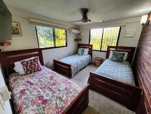 A room with three wooden beds, each with a different floral bedspread. There are windows providing natural light and views of greenery outside.