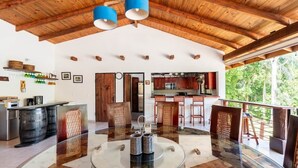 A spacious room with a glass table, wooden ceiling beams, and a kitchen area visible through an open door.