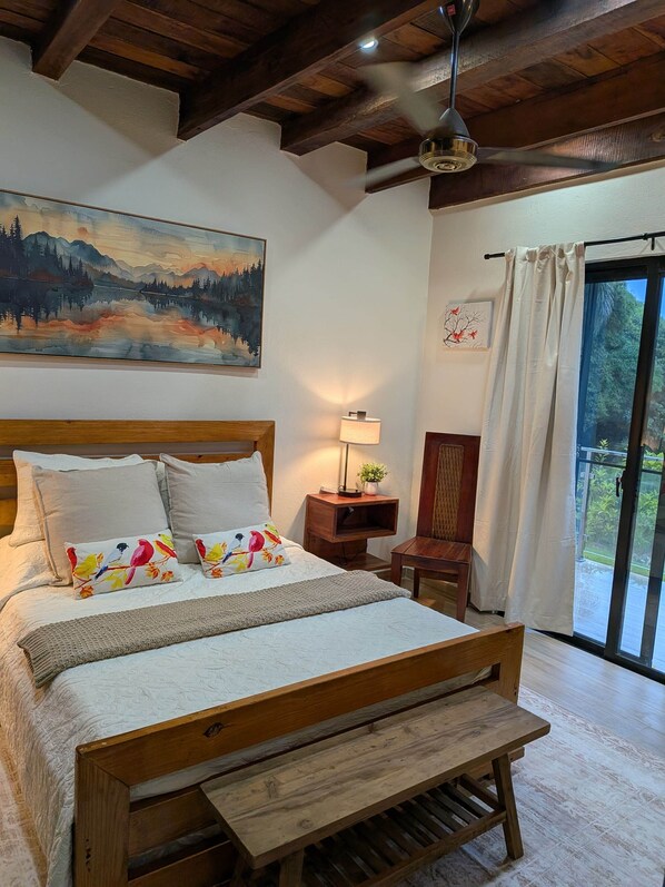 A bedroom with a wooden bed, a painting of a landscape, a nightstand with a lamp, and a view of greenery through a window.